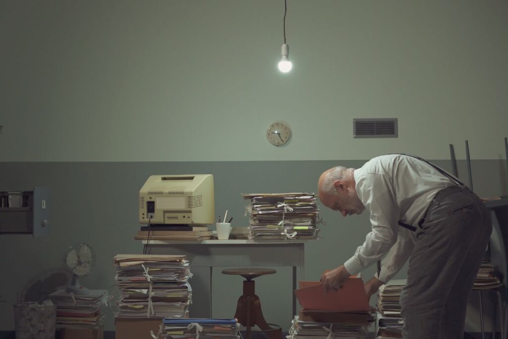 Businessman working in a messy office, he is surrounded by piles of paperwork and searching for a file