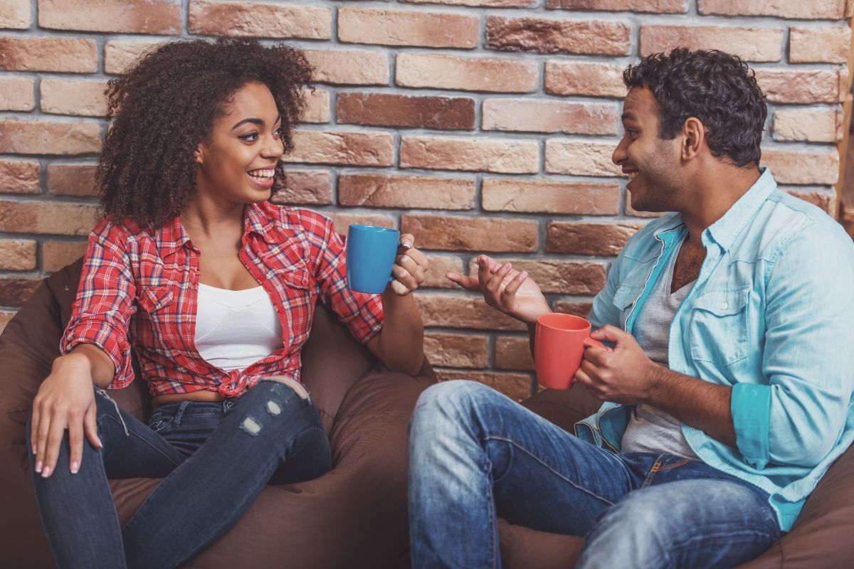 Attractive Afro-American couple drinking coffee, talking and laughing while sitting on beanbag chair against brick wall