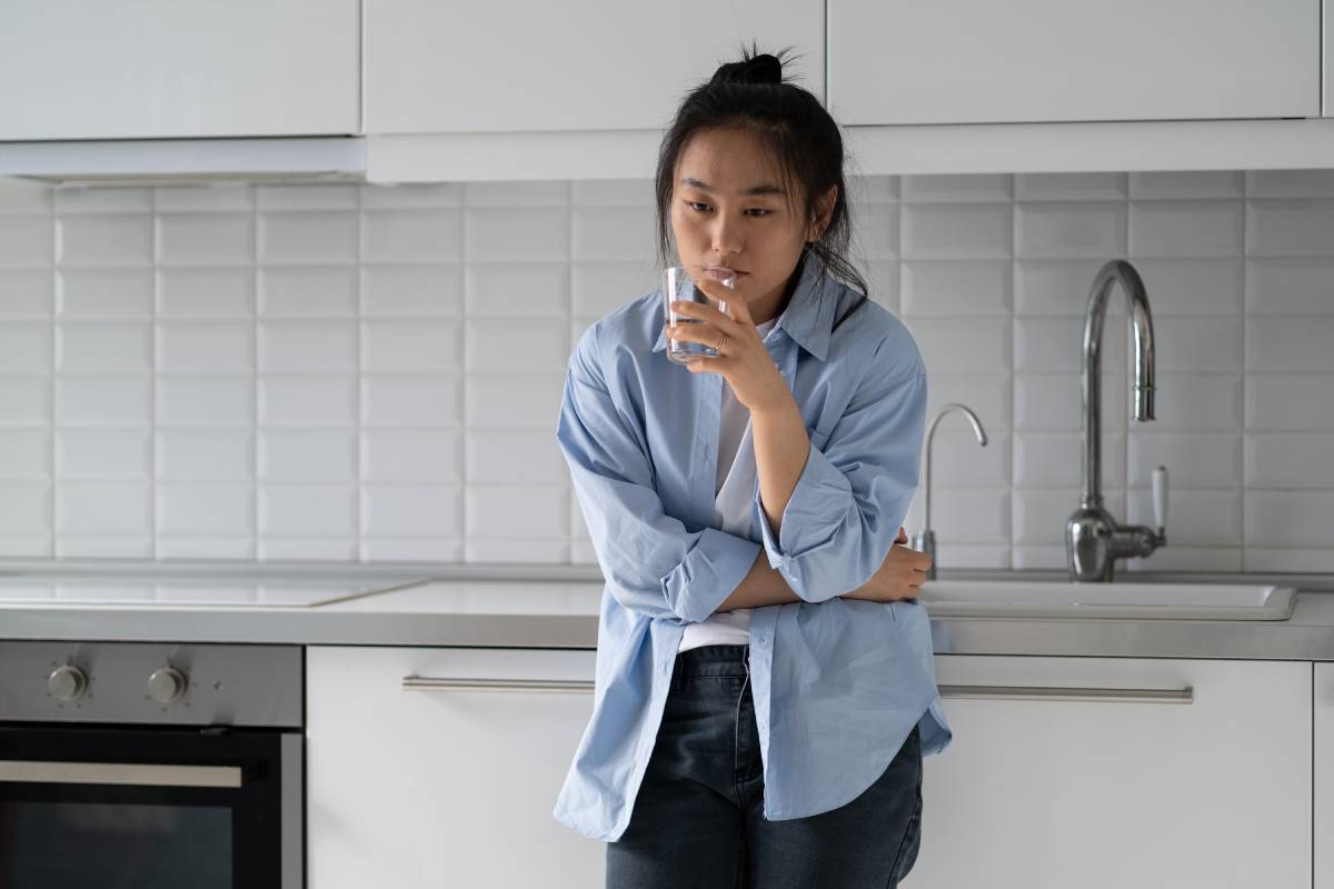 Tired unhealthy Asian woman standing in kitchen at home holding glass of water, suffering from strong sudden abdominal throbbing pain, feeling morning pain, pancreatitis gastritis from unbalanced diet