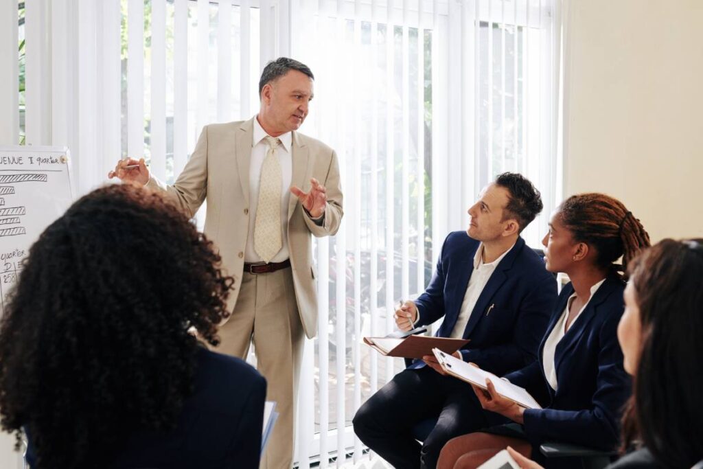 Serious mature entrerpreneur answering questions of young colleagues at meeting