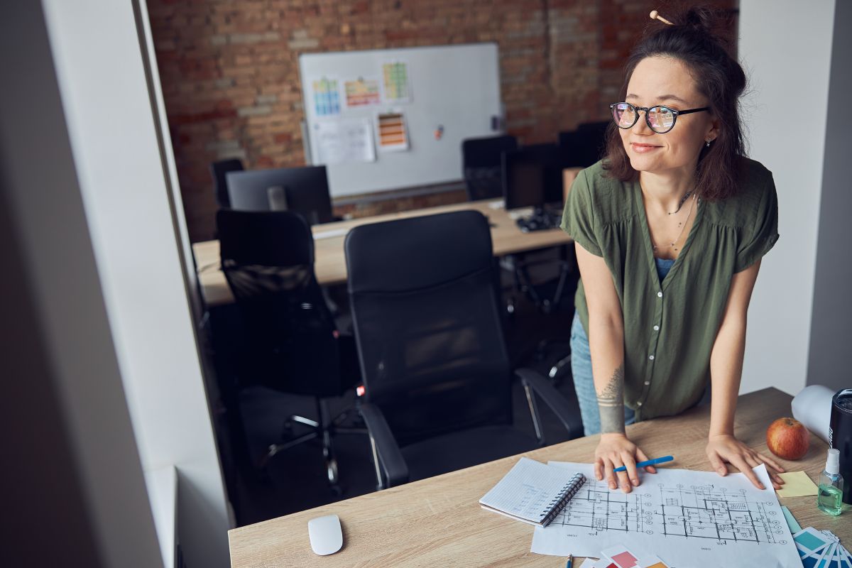 Interior designer or architect in casual wear with messy hairdo smiling aside while working on a blueprint for new project, standing in her office on a daytime. Home renovation concept