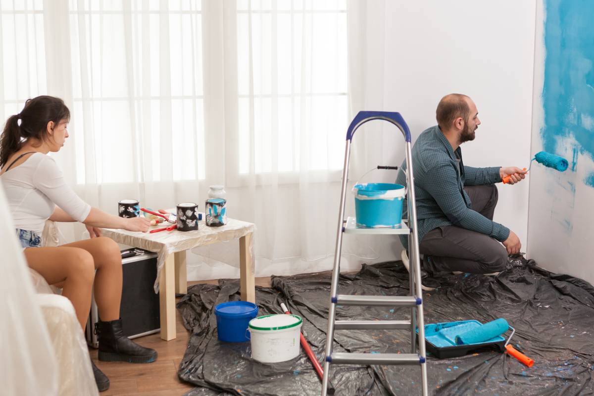 Wife resting while the husband painting the wall with roller brush. Home during renovation, decoration and painting. Interior apartment improvement maintenance. Roller, ladder for house repair.